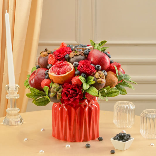 Decorative arrangement of fruits and flowers in a red vase on a table with a candle and small bowl in the background.