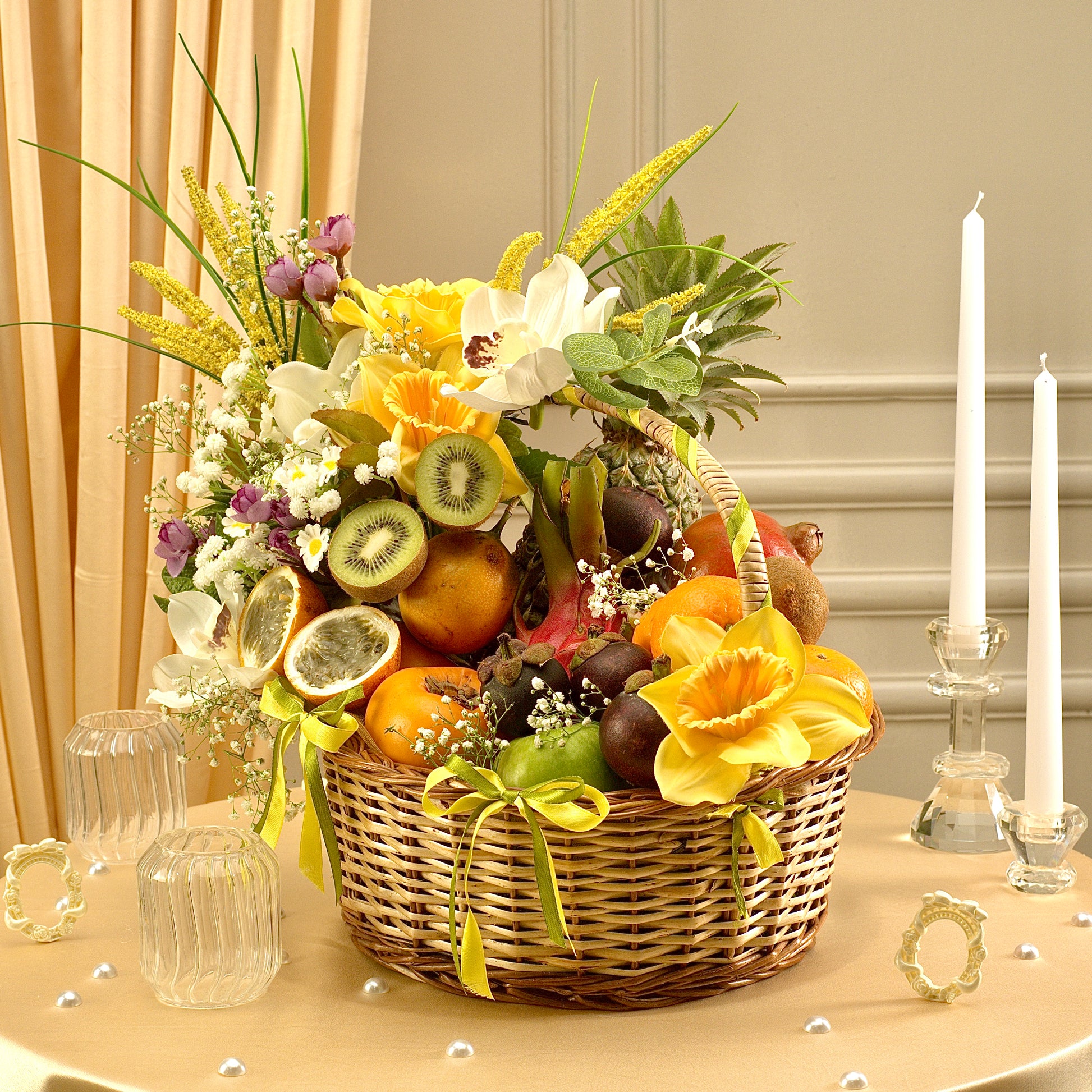 Decorative fruit and flower basket on a table with candles and glassware.