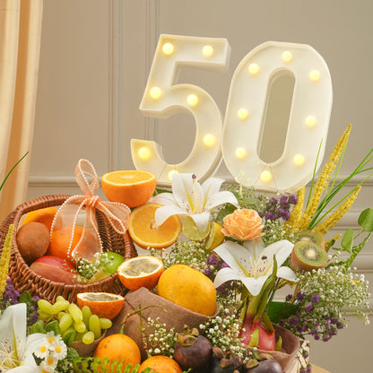 Decorative setup with fruit basket, flowers, and illuminated number '50' on a neutral background
