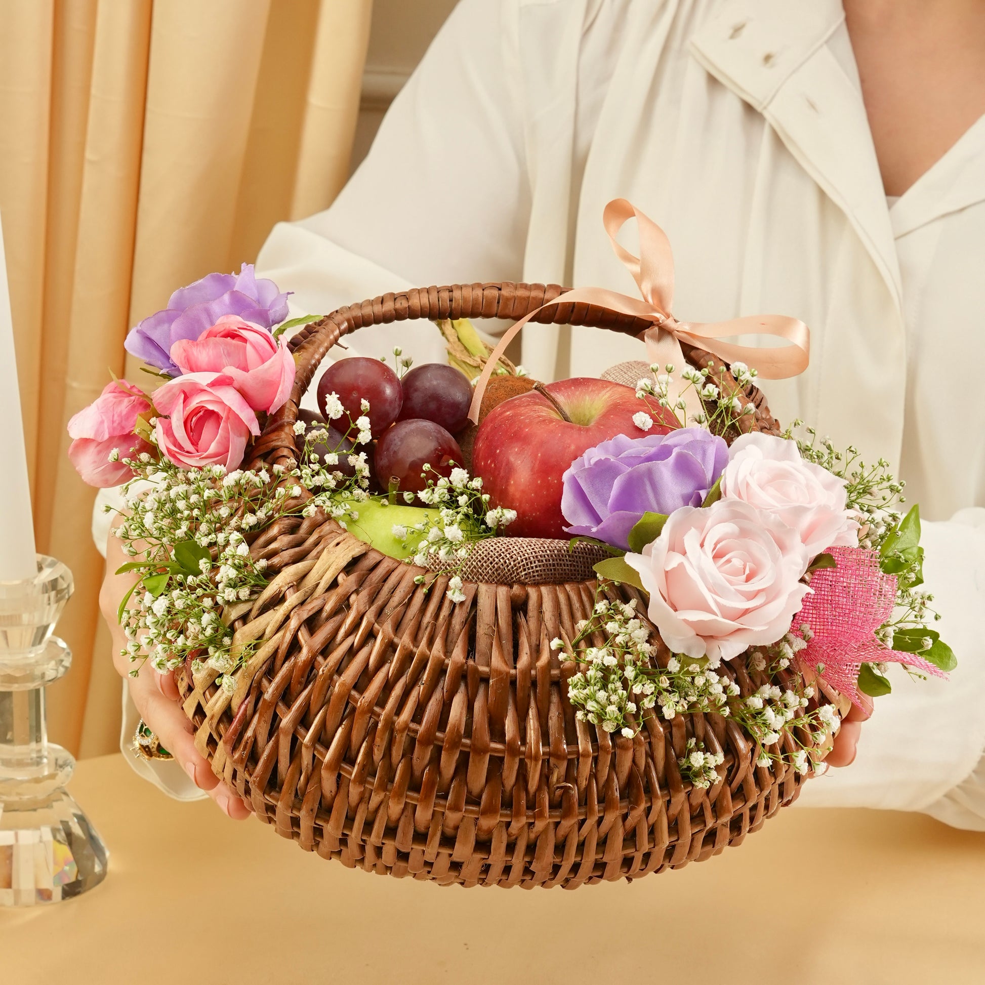 Person holding a wicker basket with fruits and flowers on a beige background