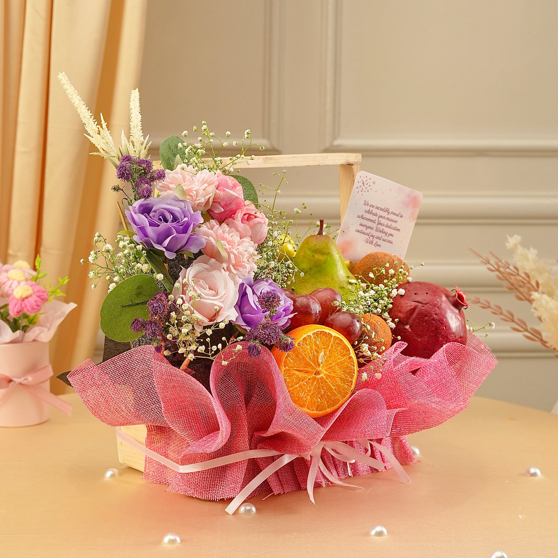 Decorative fruit and flower arrangement with pink ribbon on a table.